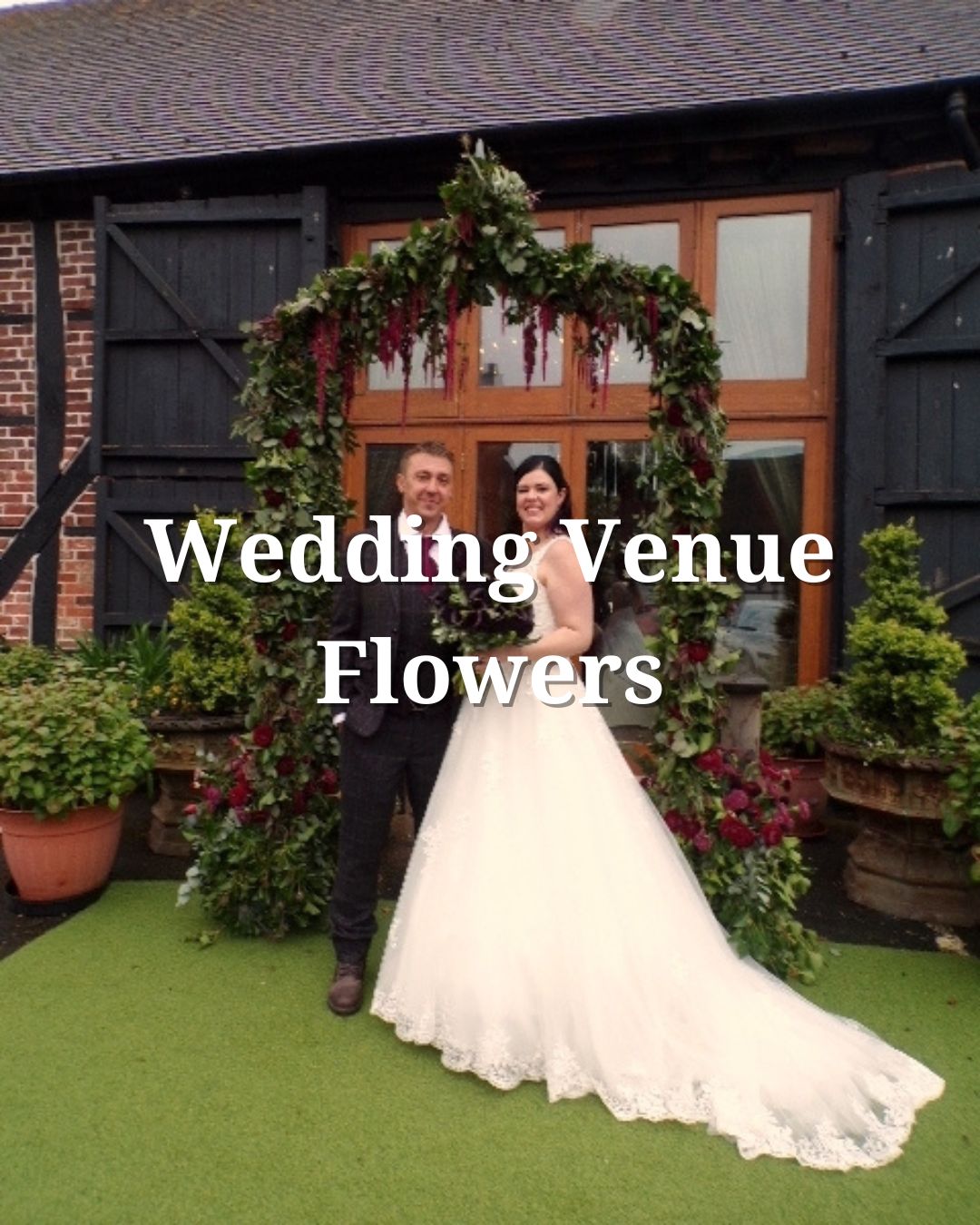 A smiling couple stands under a lush floral arch, with greenery and burgundy flowers, in front of a rustic building